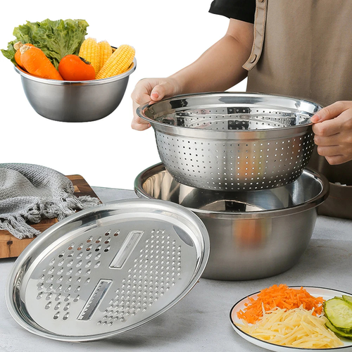 A stainless steel kitchen utensil set being demonstrated in use, including a large metal mixing bowl, a perforated steaming/colleting basket, a round grater plate, and a bowl of chopped vegetables. A person's hands and torso are visible holding the basket over the bowl, with a white studio background and food props around the set.

