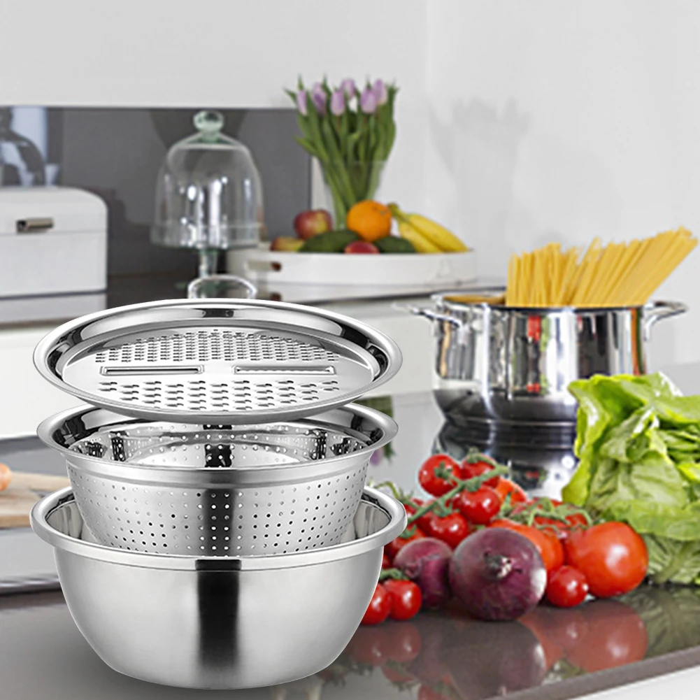 A set of stainless steel kitchen utensils, including a deep mixing bowl, a perforated steamer lid, and a matching colander basket, displayed on a kitchen countertop with fresh vegetables and cookware in the background.

